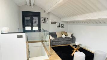 A modern living room with a grey sofa, a small round table, and a white ottoman. There is a black and white rug, a white cabinet, and a black-framed window.