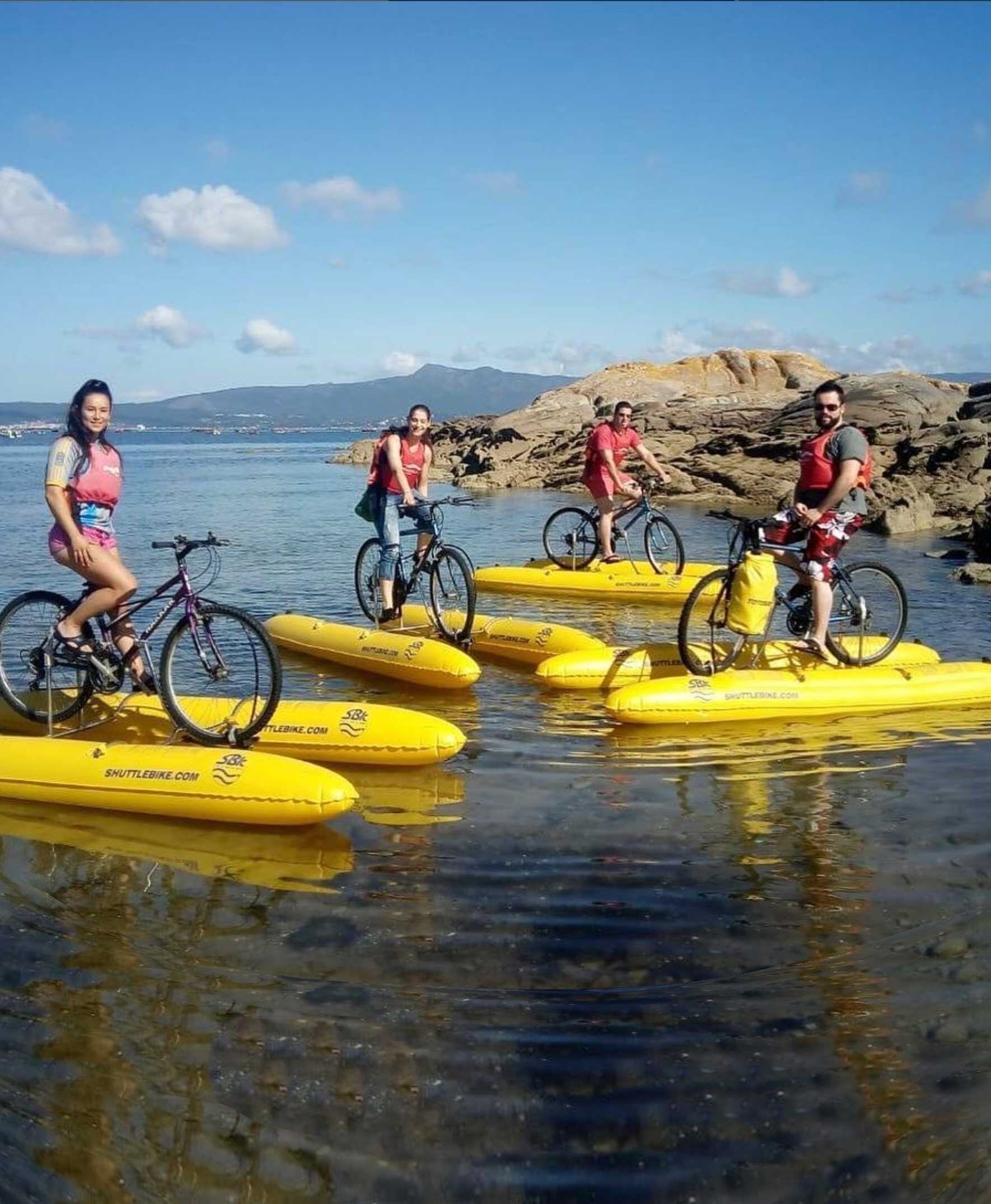 Personer cyklar på gula vattenfarkoster på en vattenyta med ett kuperat landskap och berg i bakgrunden.