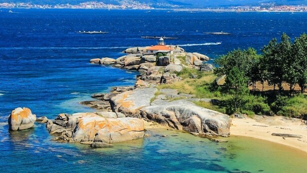 Un paisaje costero con un faro sobre un promontorio rocoso, aguas azules cristalinas y una ciudad distante en el horizonte.
