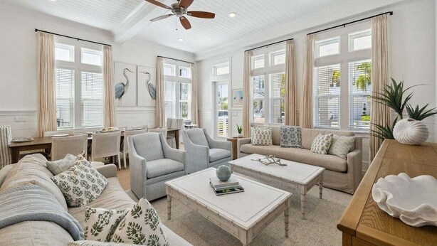 Un salon spacieux avec un plafond haut, de grandes fenêtres dotées de rideaux, un ventilateur de plafond et divers éléments de mobilier, notamment un canapé, des fauteuils et une table basse.