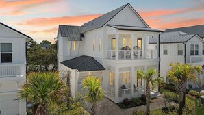 Une maison blanche à deux étages, dotée d’un balcon, entourée de palmiers et d’un jardin bien entretenu.