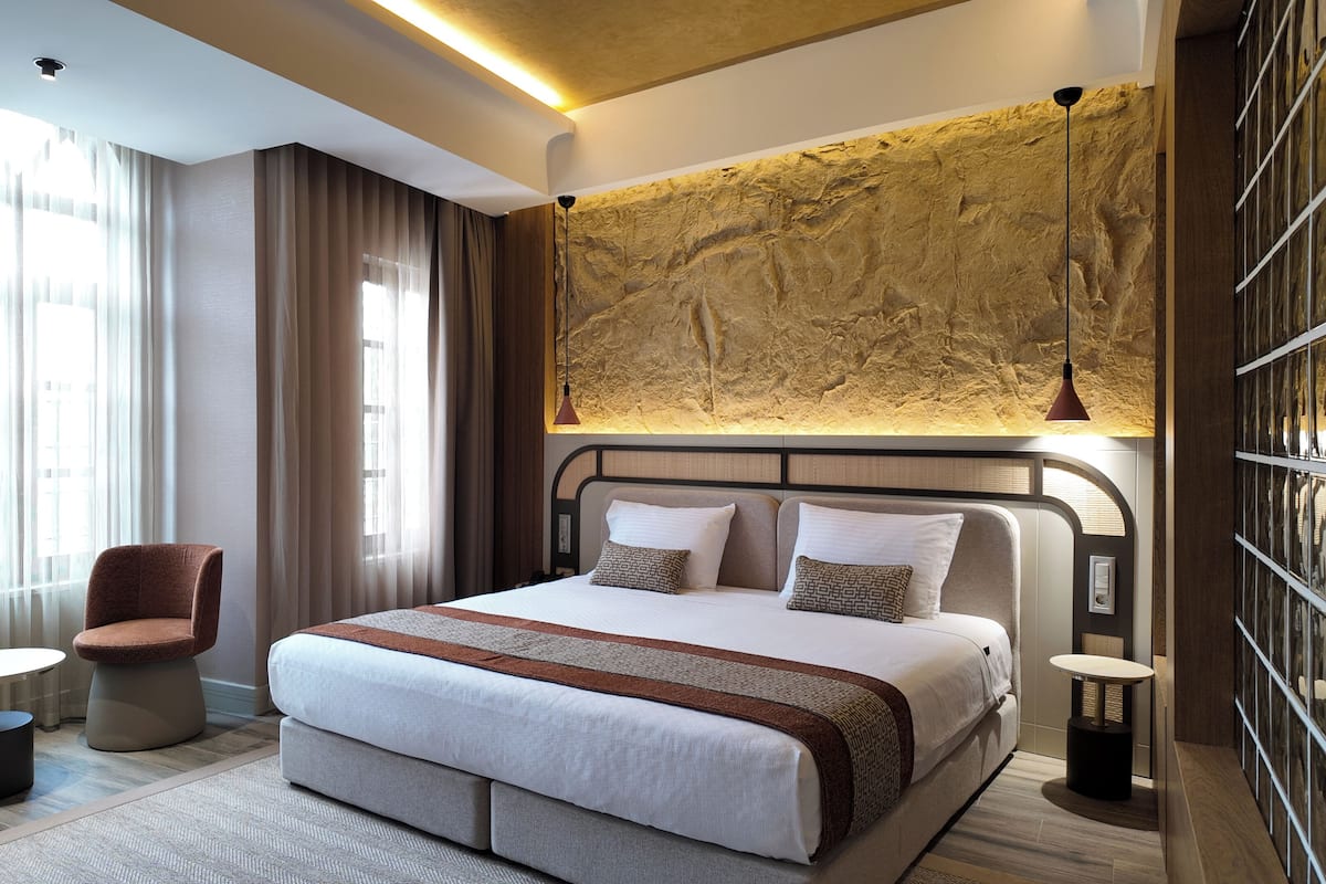 A modern hotel room with a large bed, a seating area with a chair and table, and a textured wall feature.