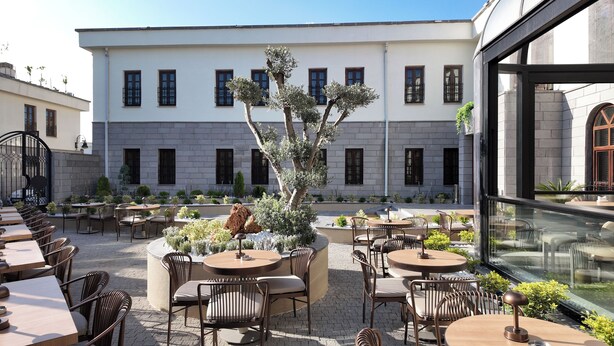 An outdoor seating area with tables and chairs, a central tree, and a building with multiple windows in the background. - Rempart Hotel (Diyarbakir)