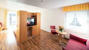 Habitación de hotel con piso de madera, un televisor en un mueble de madera, una mesa de comedor con sillas, un sofá rojo y una ventana con vista a la Torre Eiffel.