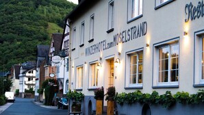 Un edificio de hotel con un cartel que dice 'WINZERHOTEL zum MOSELSTRAND' y una vista de la calle con coches aparcados y bancos.