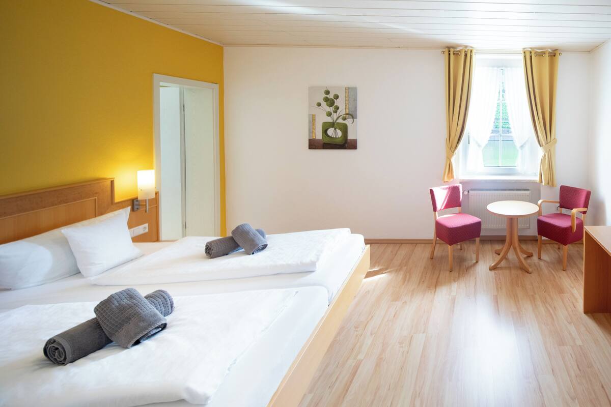 Een hotelkamer met twee bedden, een tafeltje, twee stoelen, een raam met gordijnen en een schilderij aan de muur.