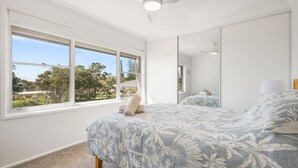 Een slaapkamer met een groot raam, een bed met bloemenbedrukking, een plafondventilator en een spiegel.