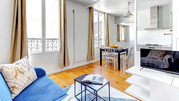 A modern living room with a blue sofa, a dining area with a table and chairs, a kitchen with a stove and cabinets, and large windows with curtains. - Cozy 1BR Near Champs-Élysées - Sleeps 4 (Paris)