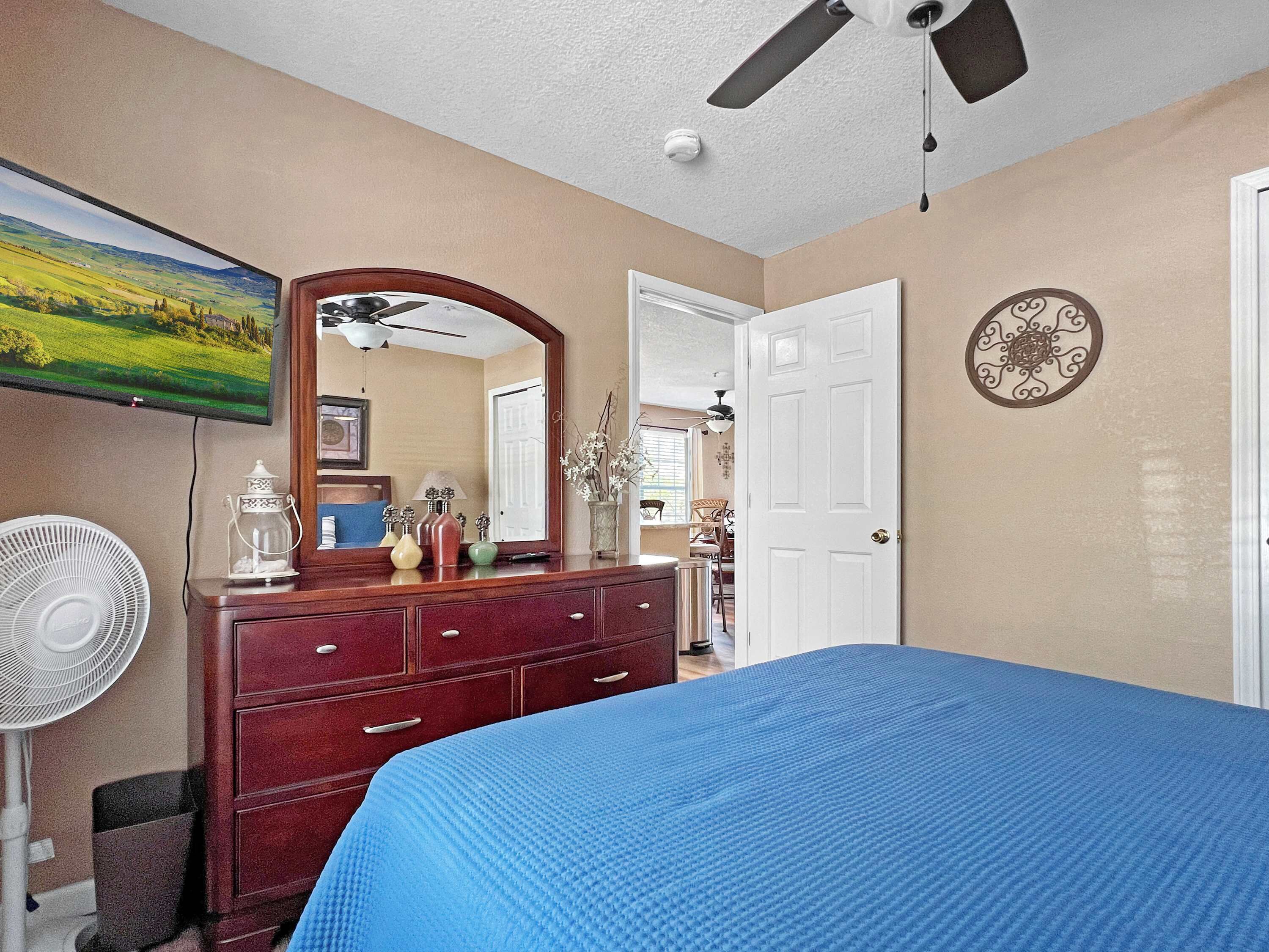 A bedroom with a bed, dresser, mirror, fan, and a TV on the wall.