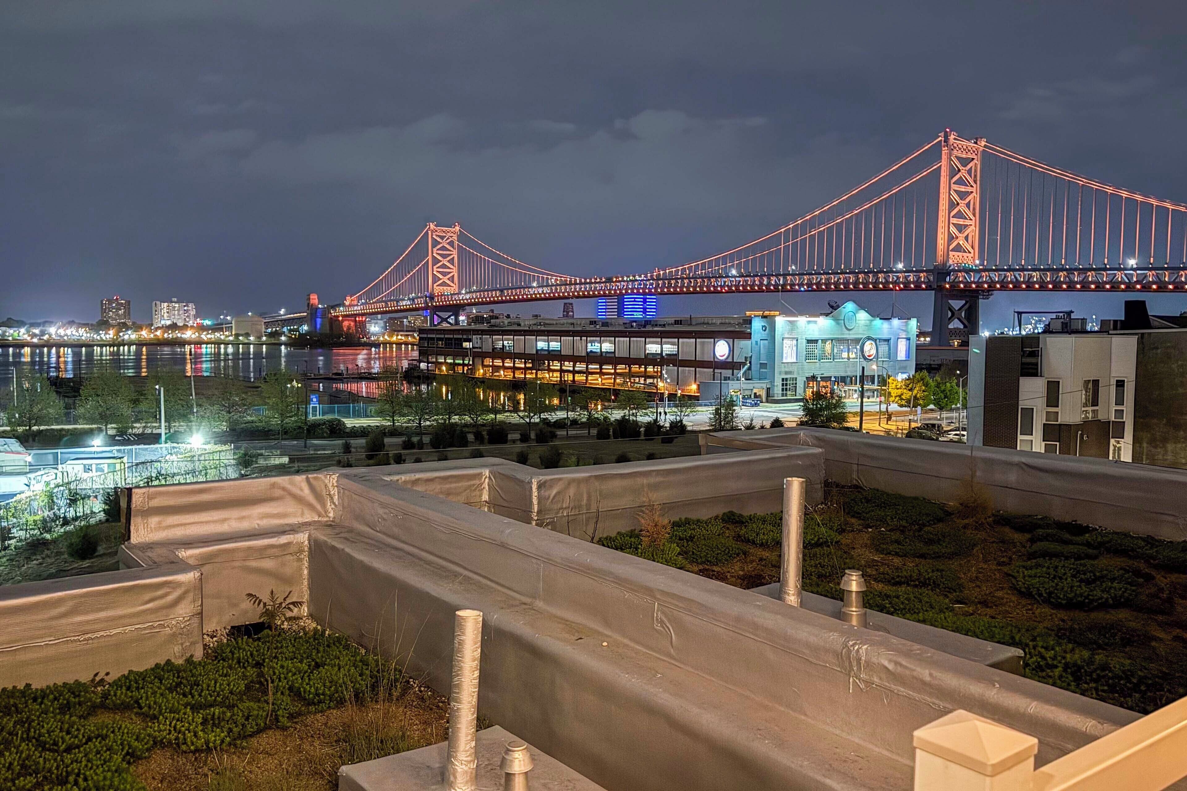 A nighttime view of a bridge with illuminated cables, a modern building with blue lighting, and a landscaped area with concrete planters.