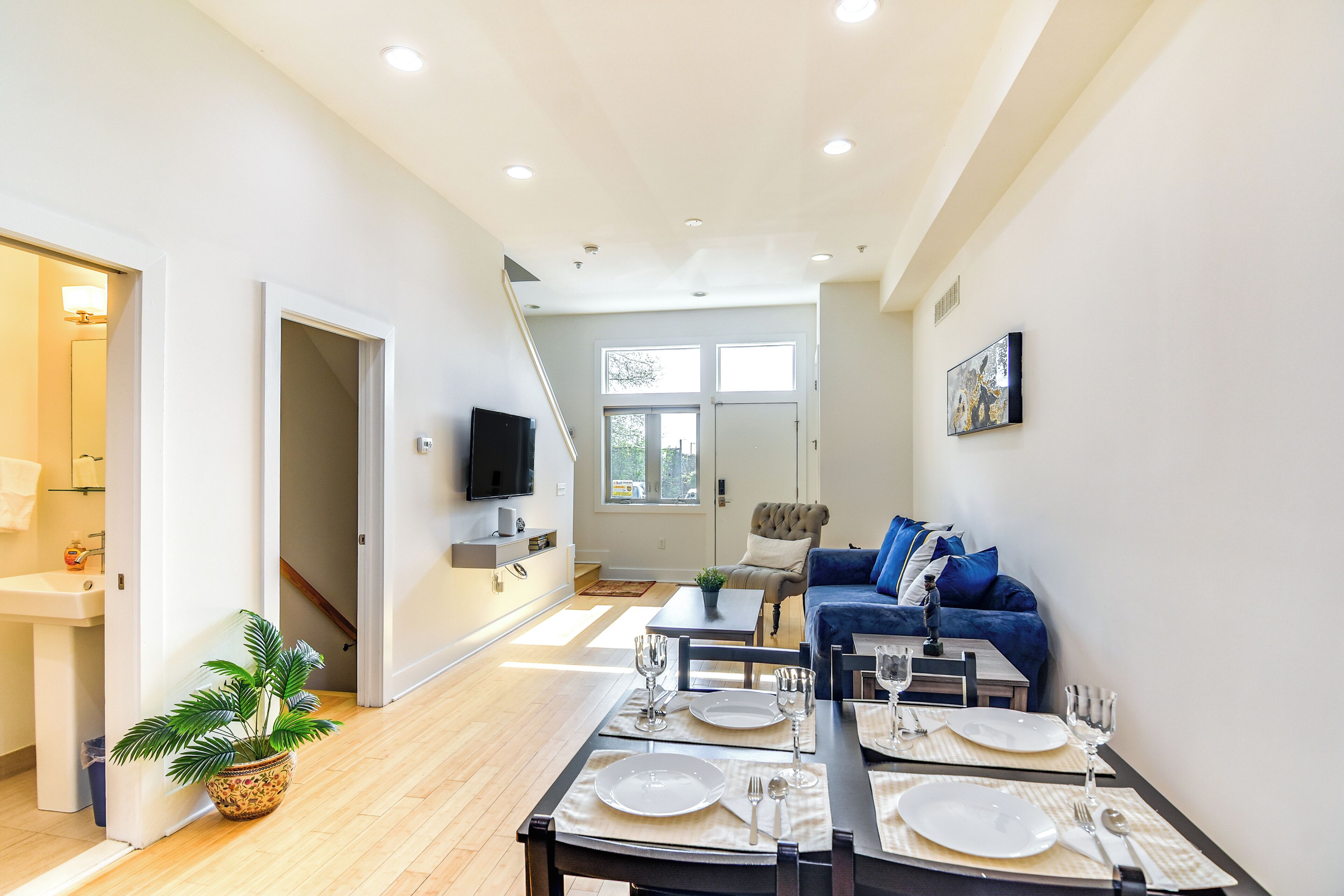 A modern living room with a dining area set for six, a sofa, a TV, and a staircase leading to another room.