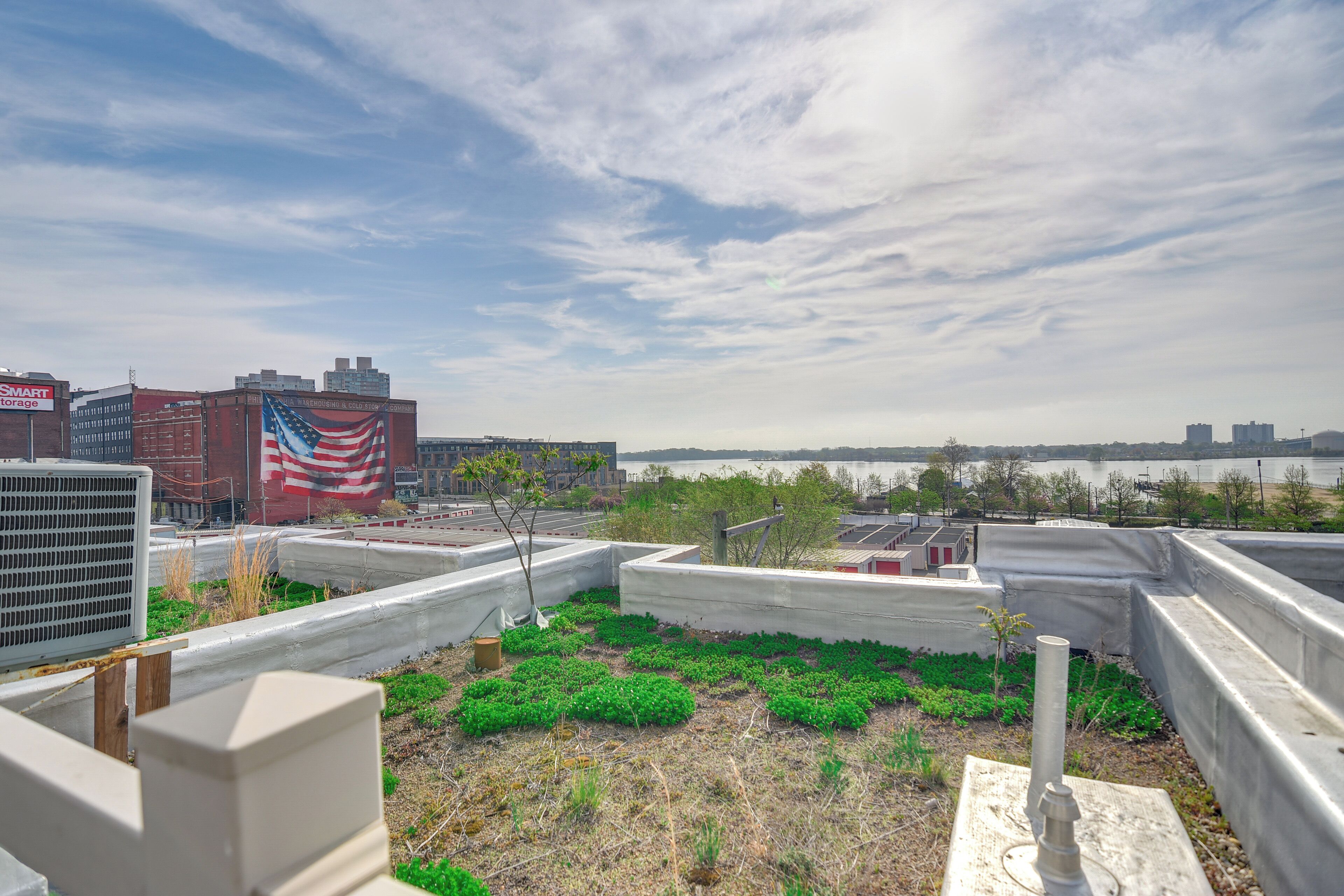 A rooftop garden with green plants, a flag, and a view of a cityscape and water body.