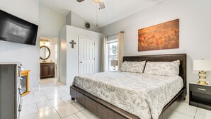Een slaapkamer met een groot bed, een aan de muur bevestigde televisie, een plafondventilator en een raam met gordijnen.