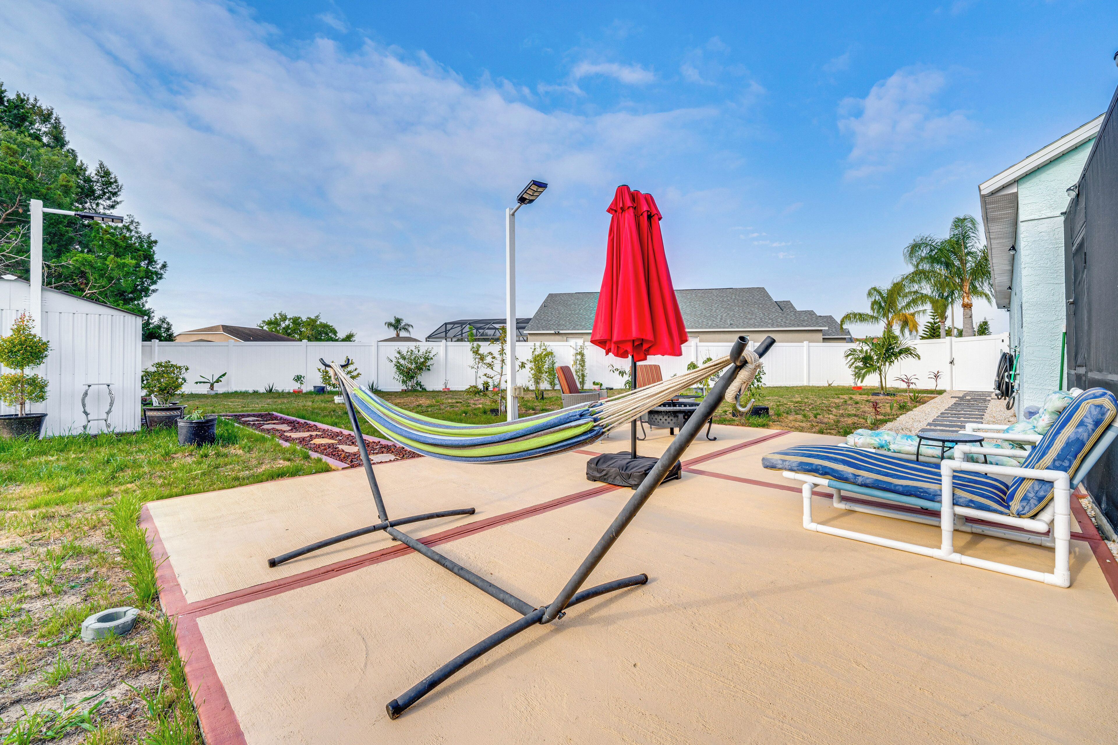 Un hamac, un parasol rouge et une chaise longue dans un jardin avec une terrasse en béton.