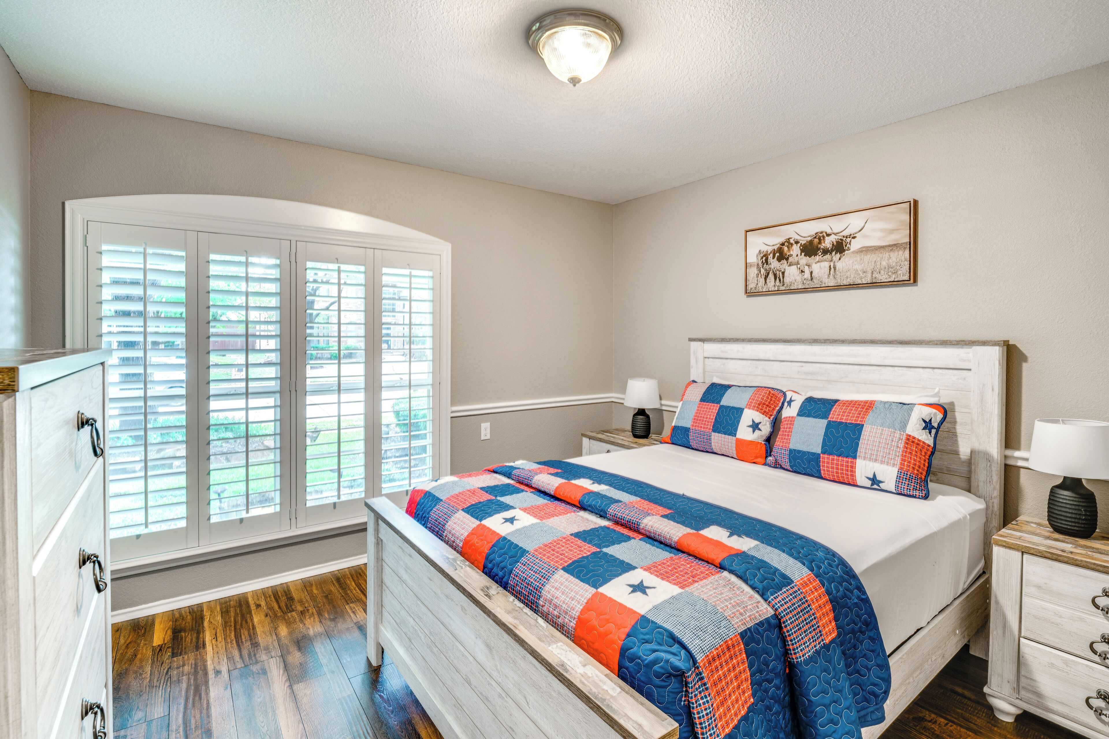 A bedroom with a wooden bed, a nightstand, a lamp, a window with blinds, and a wall-mounted picture.