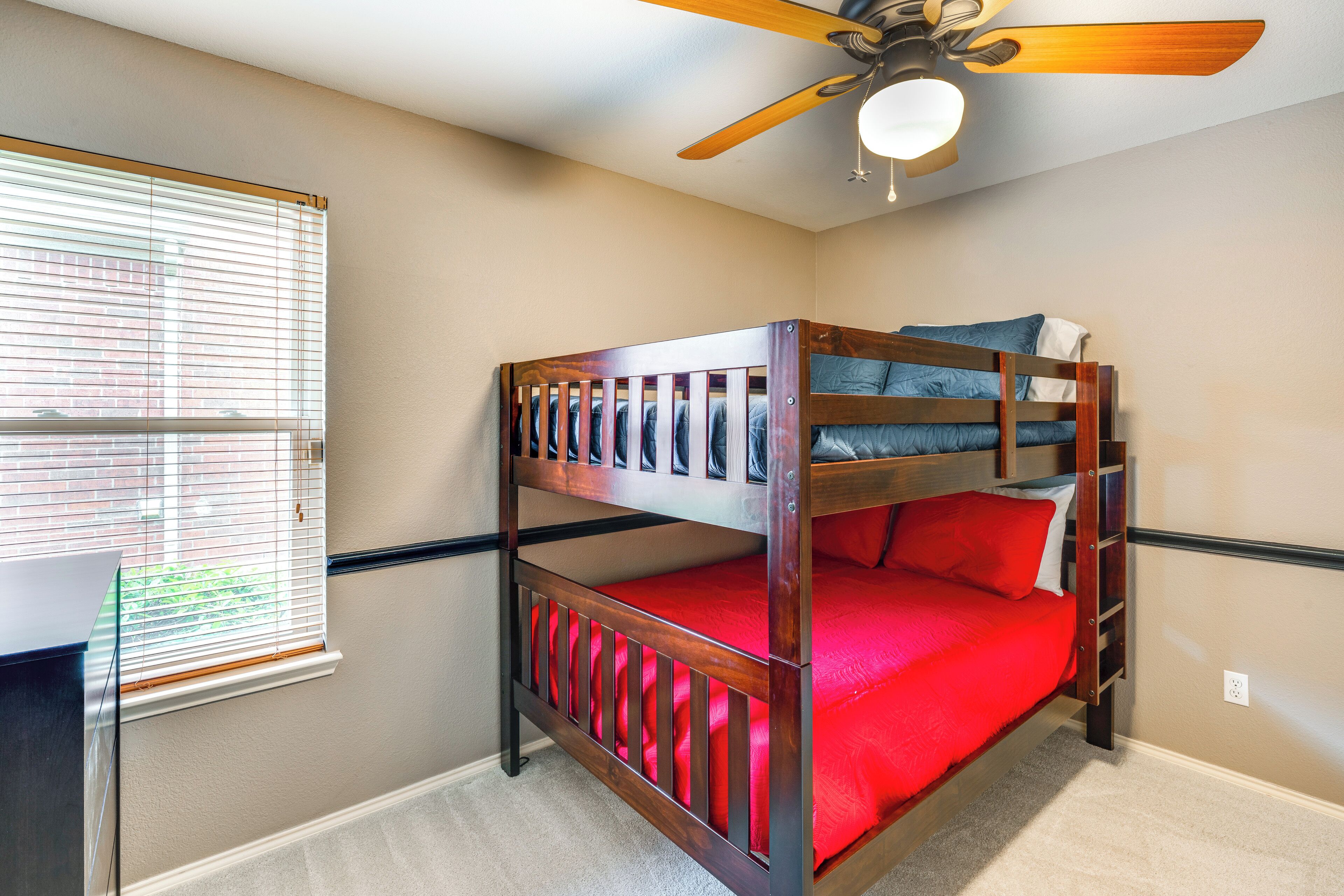 A bunk bed with a red mattress and wooden frame, a ceiling fan, and a window with blinds.