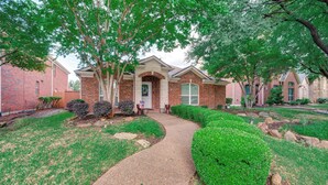 A brick house with a paved driveway, surrounded by well-maintained lawns and trees. - Swim, Game, Relax & More: Family Getaway in Frisco (Frisco)