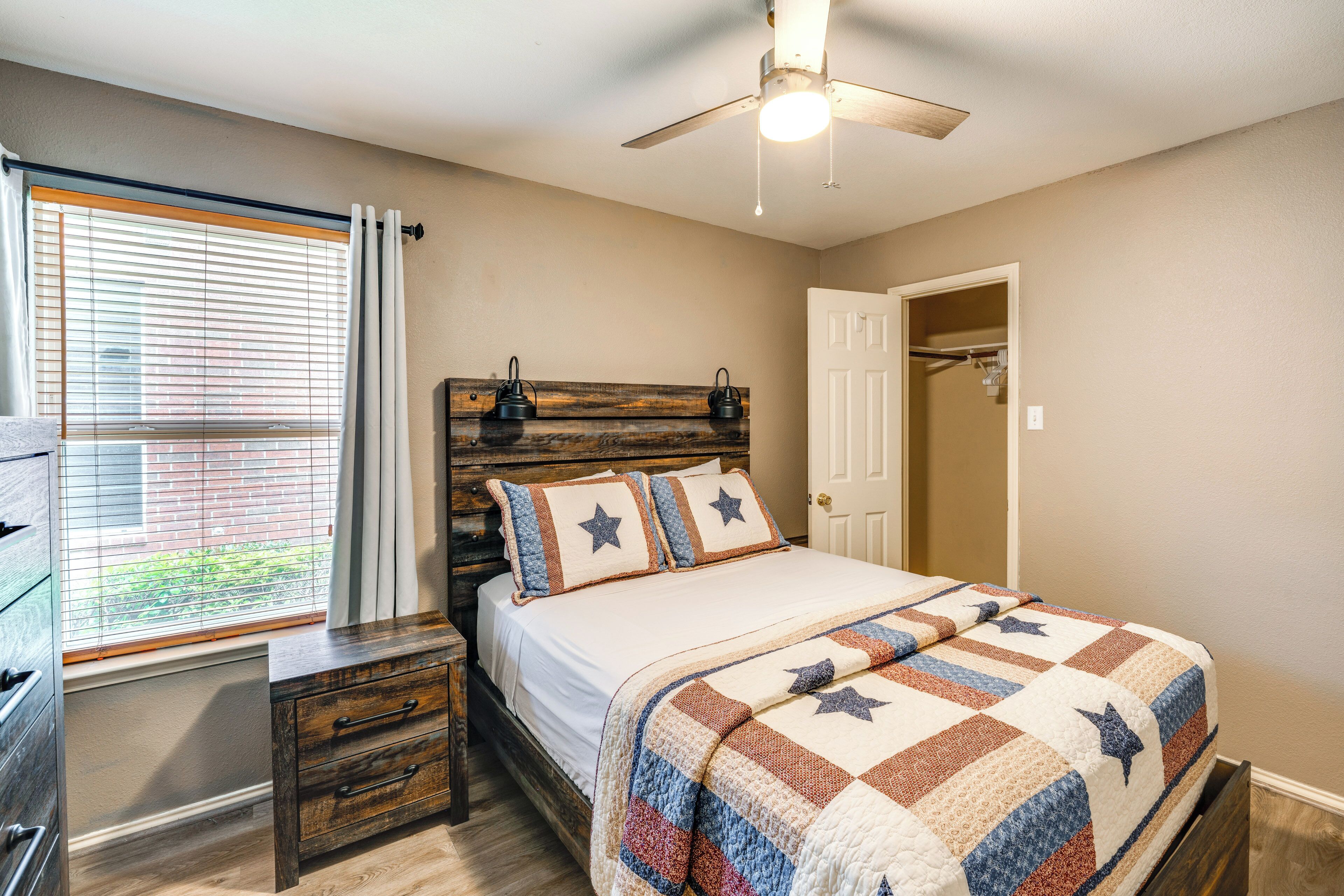 A bedroom with a wooden headboard, a bed with a quilt, a nightstand, a dresser, and a window with blinds.