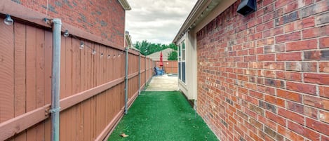 A narrow pathway flanked by brick walls, leading to a building with a visible door and windows.