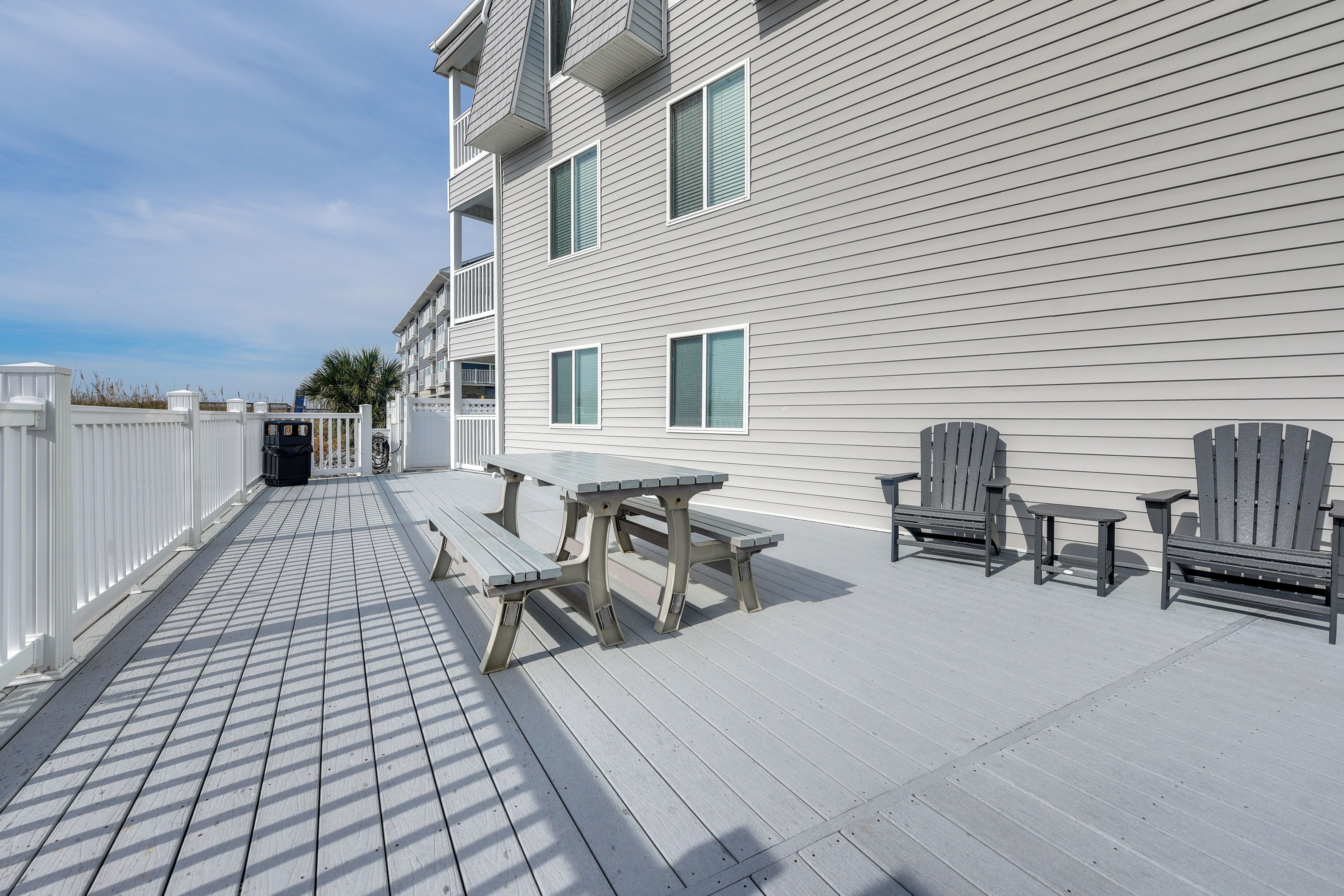 A wooden deck with picnic tables and chairs, a white fence, and a building with multiple windows.
