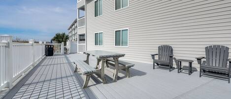 A wooden deck with picnic tables and chairs, a white fence, and a building with multiple windows.