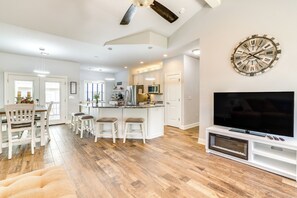 A modern kitchen with white cabinetry, a central island with stools, a large clock on the wall, and a flat-screen TV on a white stand. - < 1 Mi to Golf & Marina: Inviting Home in Afton! (Afton)