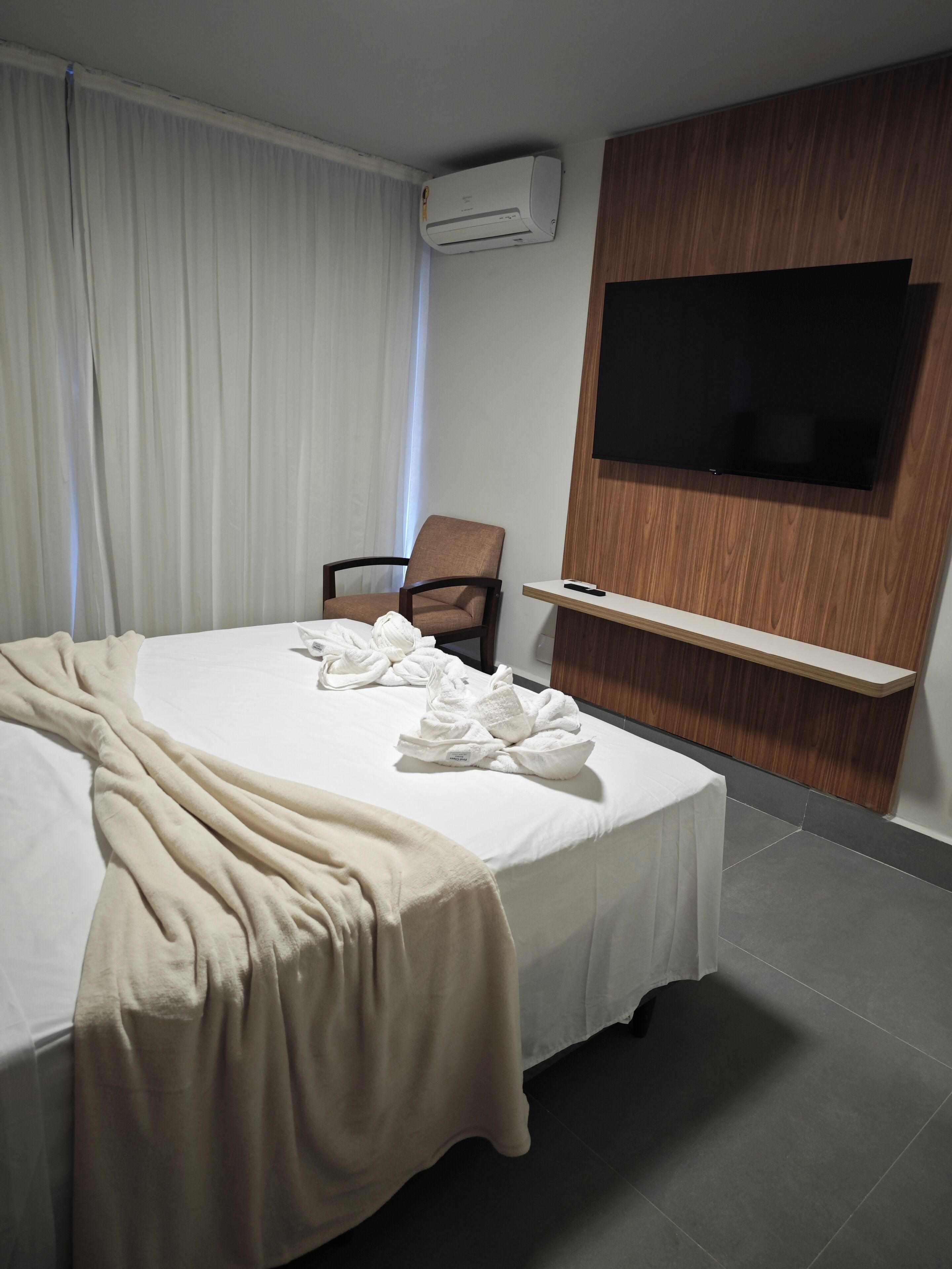 Quarto de hotel com cama, uma cadeira, televisão na parede e ar condicionado.