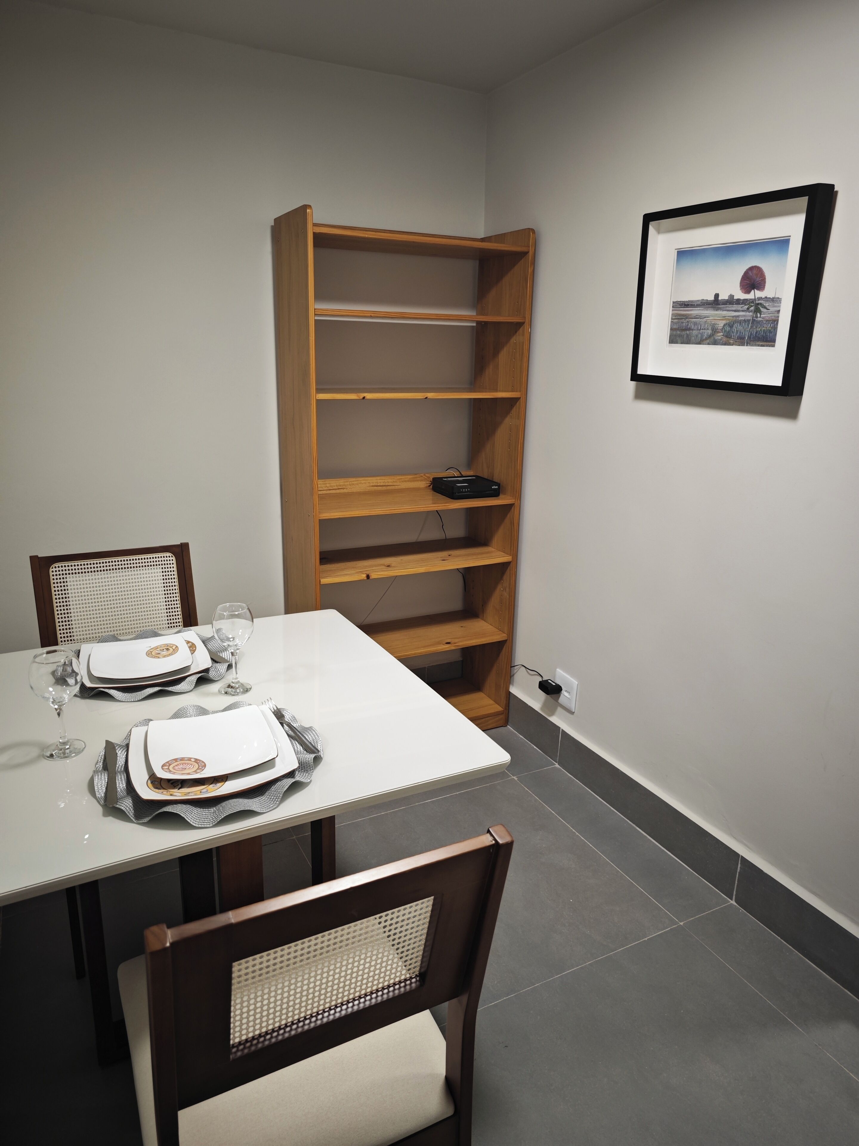 A dining area with a white table set for two, a wooden bookshelf, and a framed picture on the wall.