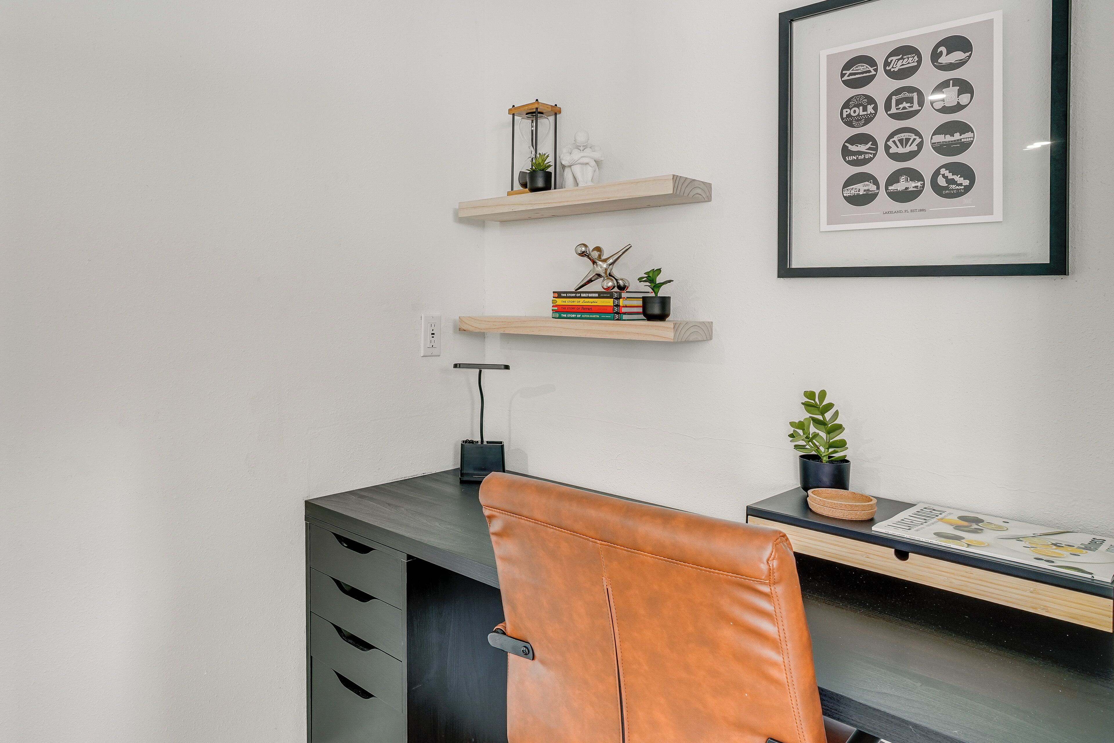 A modern office desk with a leather chair, a framed picture of logos, and a small shelf with decorative items.