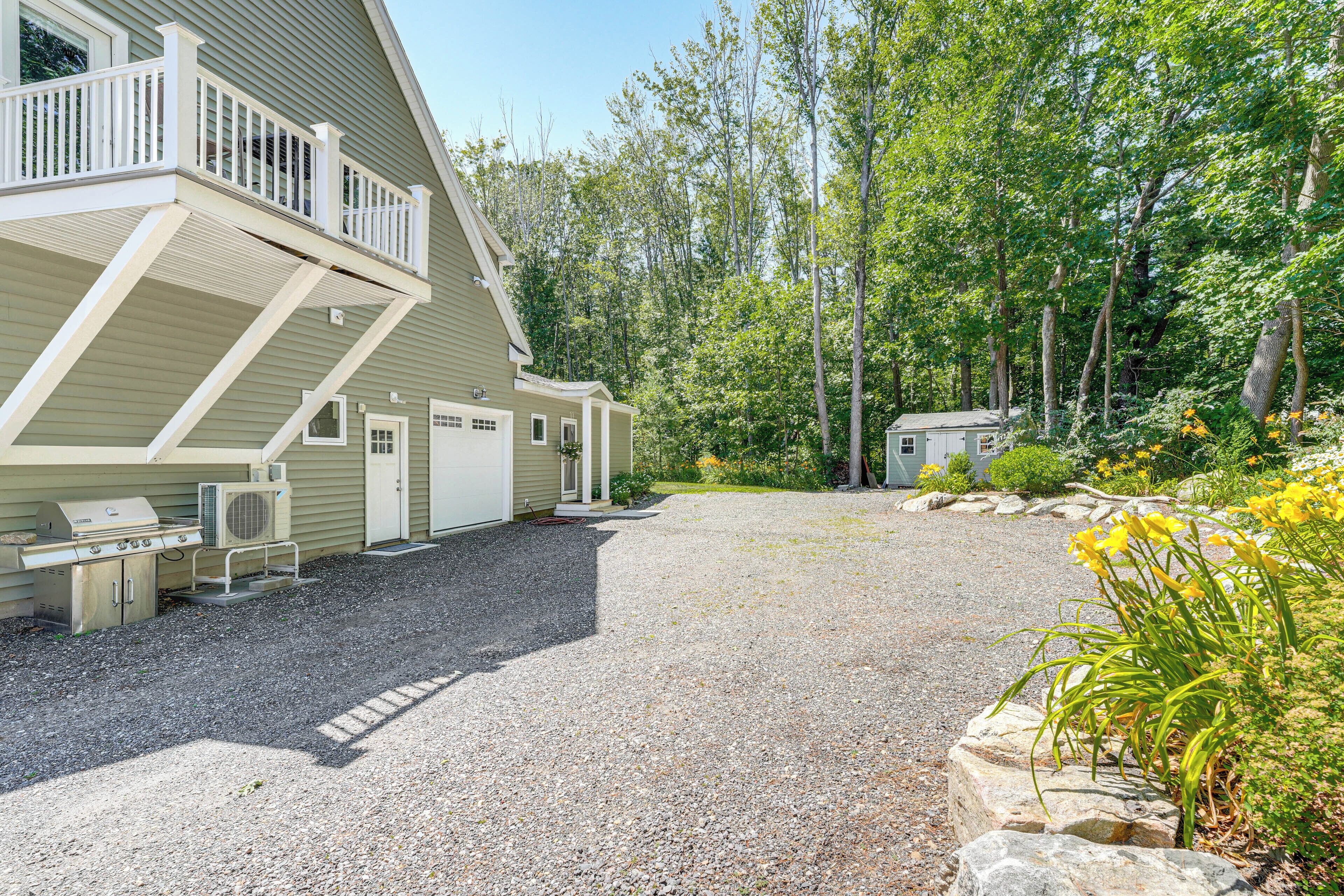 A two-story house with a gravel driveway, a barbecue area, and a small shed in a wooded area.