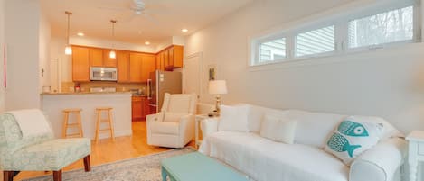 A spacious living area with a white sofa, a teal coffee table, and a patterned rug. A kitchen with wooden cabinets and a microwave is visible in the background.