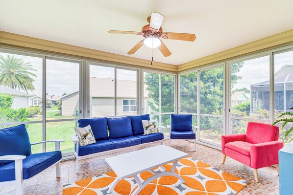 A sunroom with a ceiling fan, large windows, a blue sofa, a red chair, and a white coffee table. - Peaceful Escape w/ Golf Cart in The Villages! (The Villages)