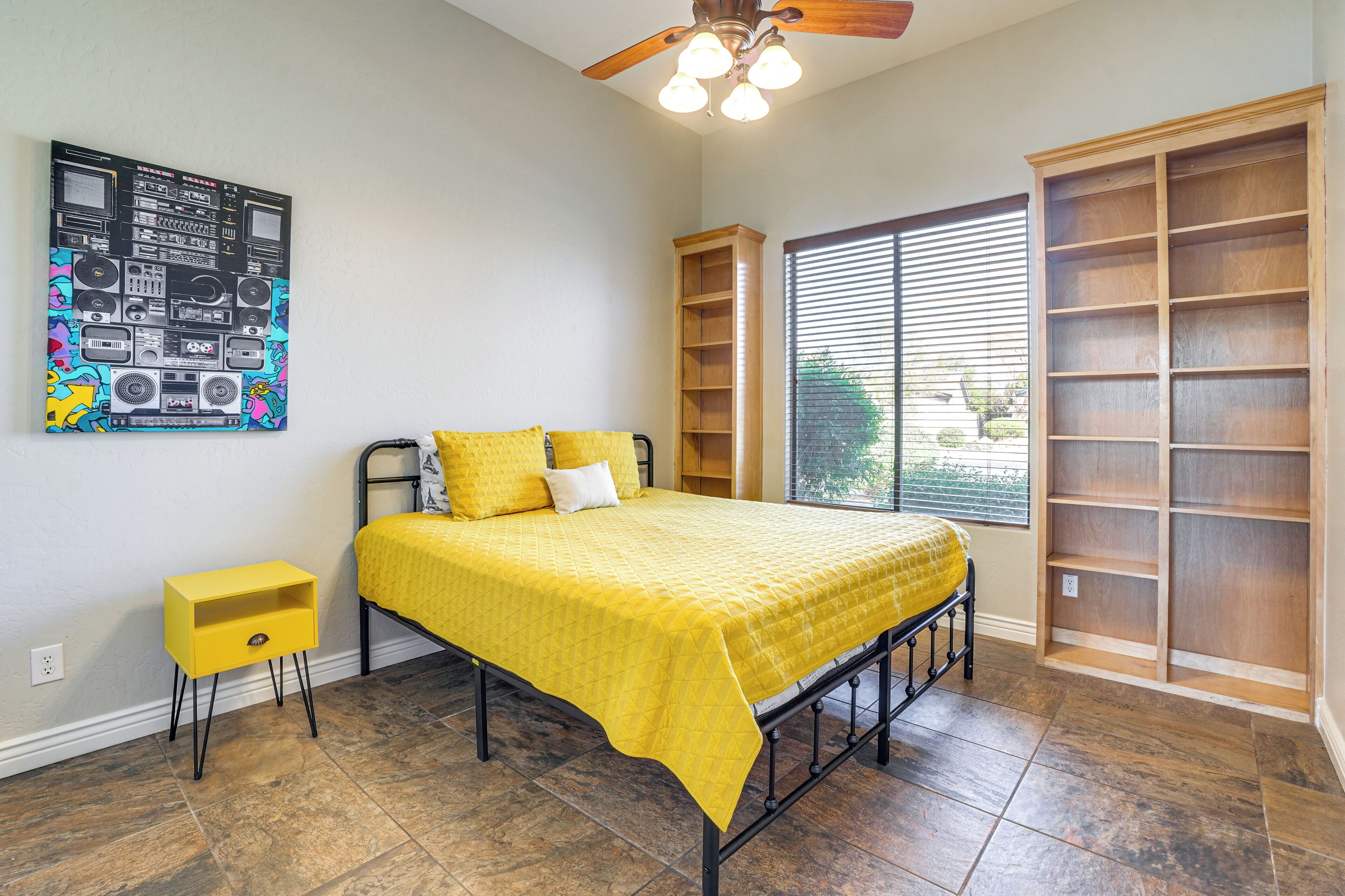 Quarto com cama, mesa de cabeceira, uma obra de arte na parede, ventoinha no teto e janela com persianas.