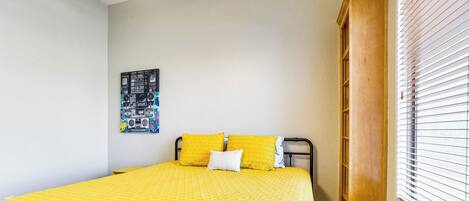 A bedroom with a bed, yellow bedding, a wall art piece, a window with blinds, and a wooden door.
