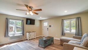Un vaste salon doté d’un plancher en bois franc, d’un ventilateur de plafond, d’un téléviseur fixé au mur et d’une grande fenêtre avec des rideaux.