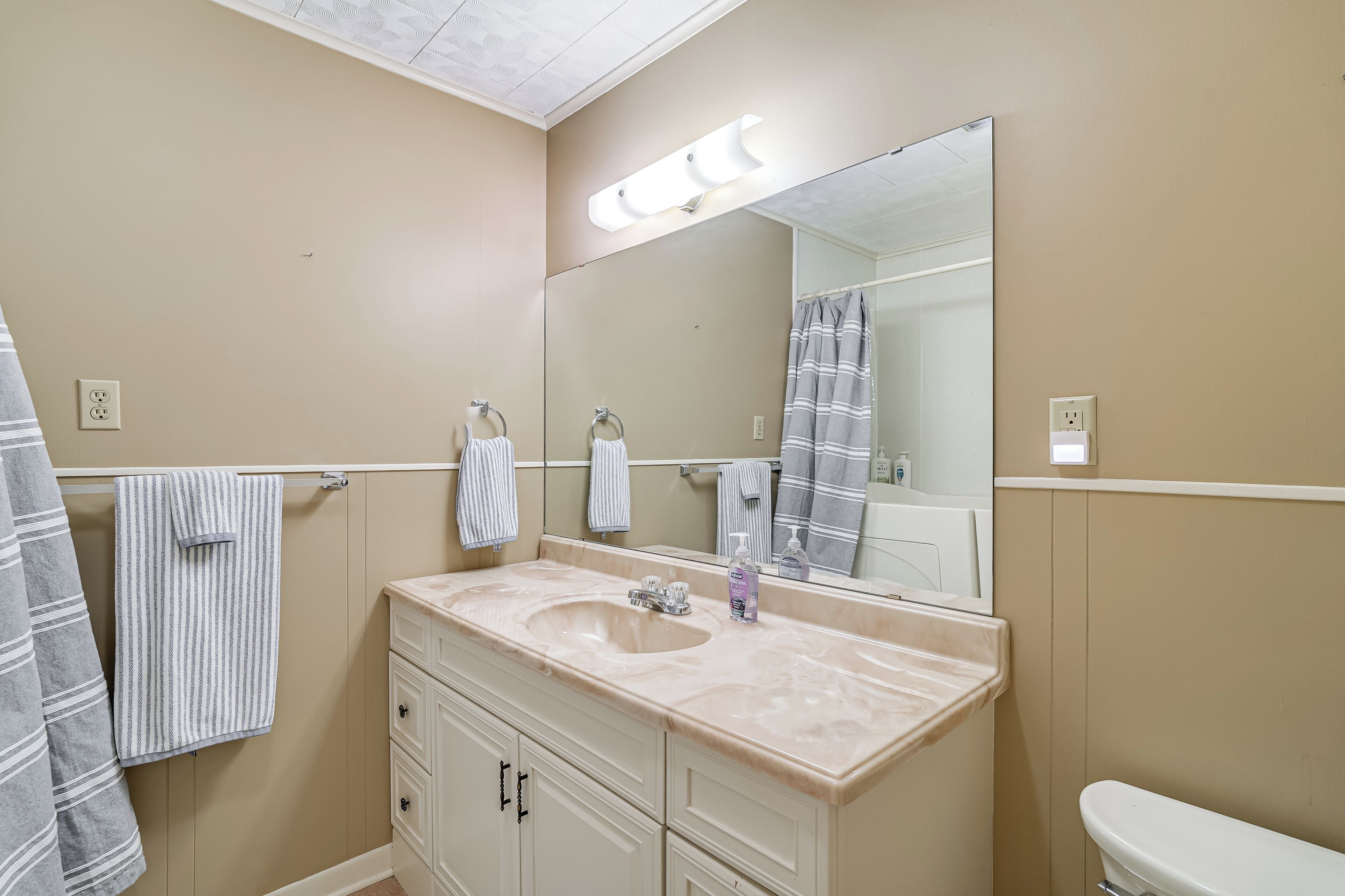 Un baño con pared color crema, un espejo grande, un lavamanos con encimera de mármol y un inodoro.