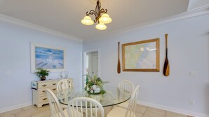 Un comedor con una mesa de vidrio rodeada de sillas blancas, una araña, un mapa en la pared y un bate de madera.