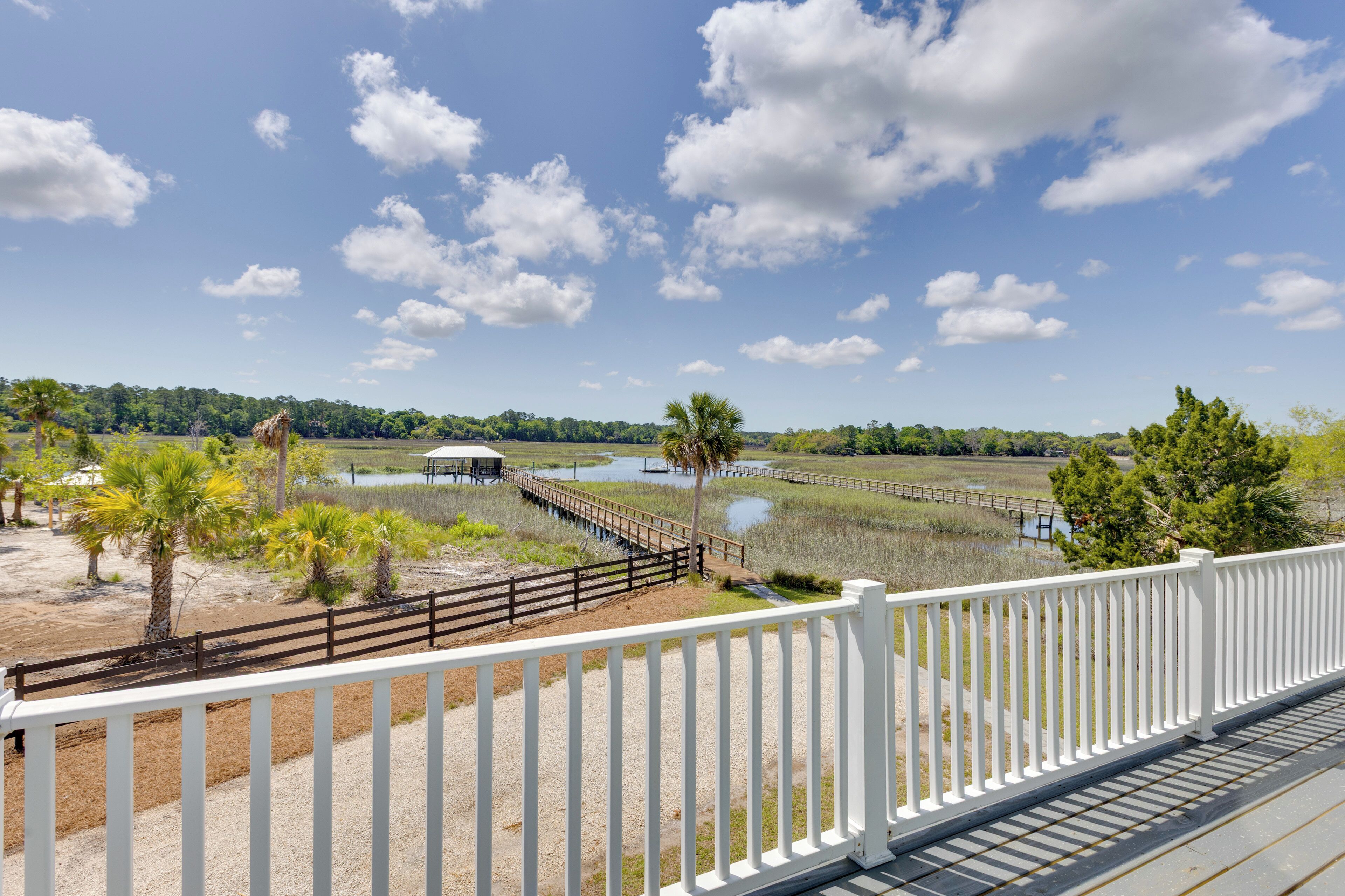 A view from a balcony with a white railing overlooking a natural landscape with water bodies, a wooden bridge, and a distant tree line.
