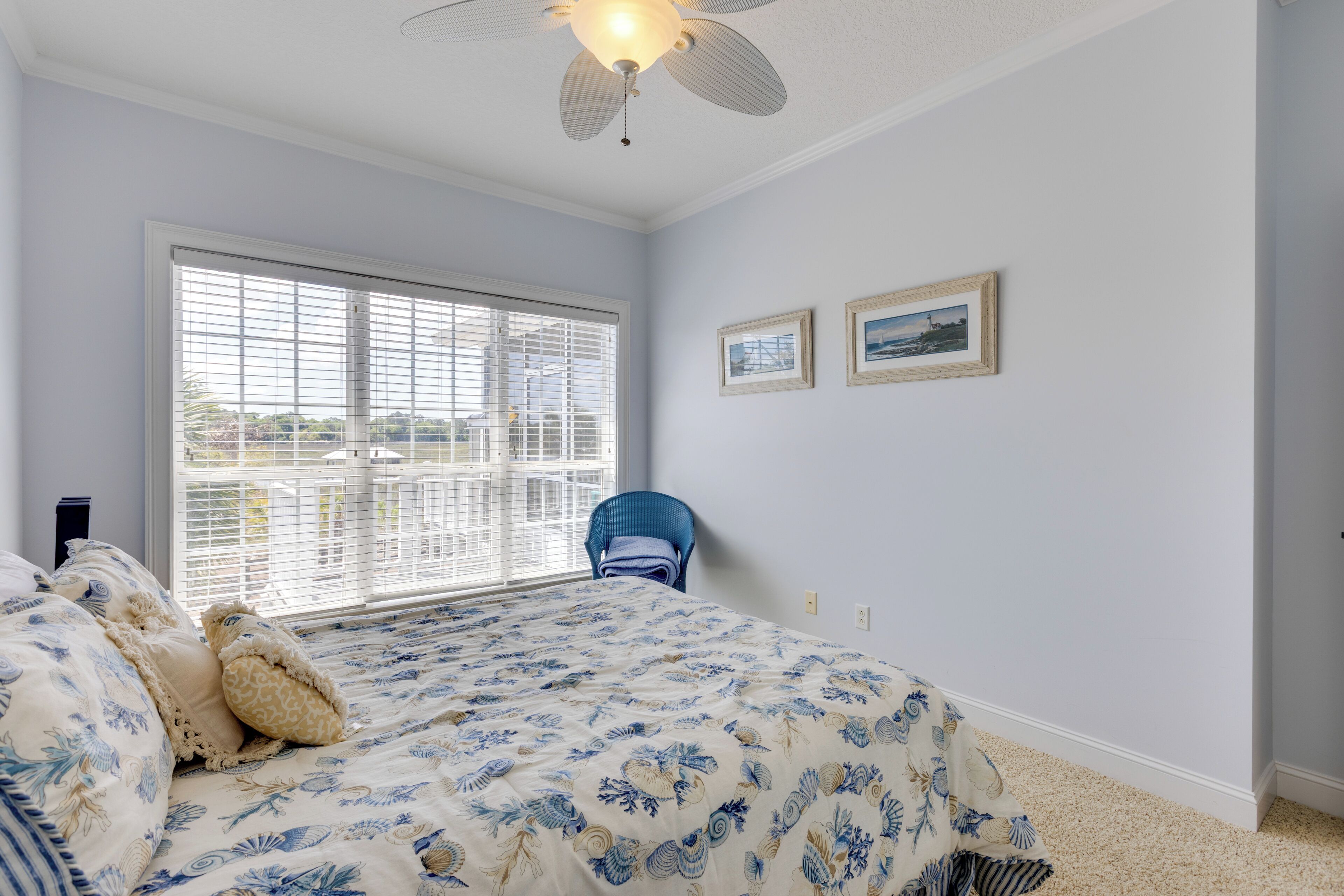 A bedroom with a large window, a bed with floral bedding, a ceiling fan, and two framed pictures on the wall.