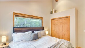 Quarto com uma cama grande, cabeceira de madeira, janela com vista para árvores, porta de madeira e mesa de cabeceira com abajur.
