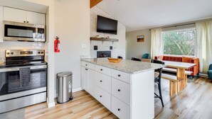 A modern kitchen with a built-in oven, microwave, and a TV mounted on a brick wall. There is a dining area with a wooden table and chairs, and a window with curtains. - Beach Access: Bbq-ready Poconos Family Getaway! (Blakeslee)