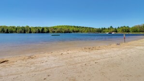 A sandy beach with a calm lake, a green boat, and a forested hill in the background. - Beach Access: Bbq-ready Poconos Family Getaway! (Blakeslee)