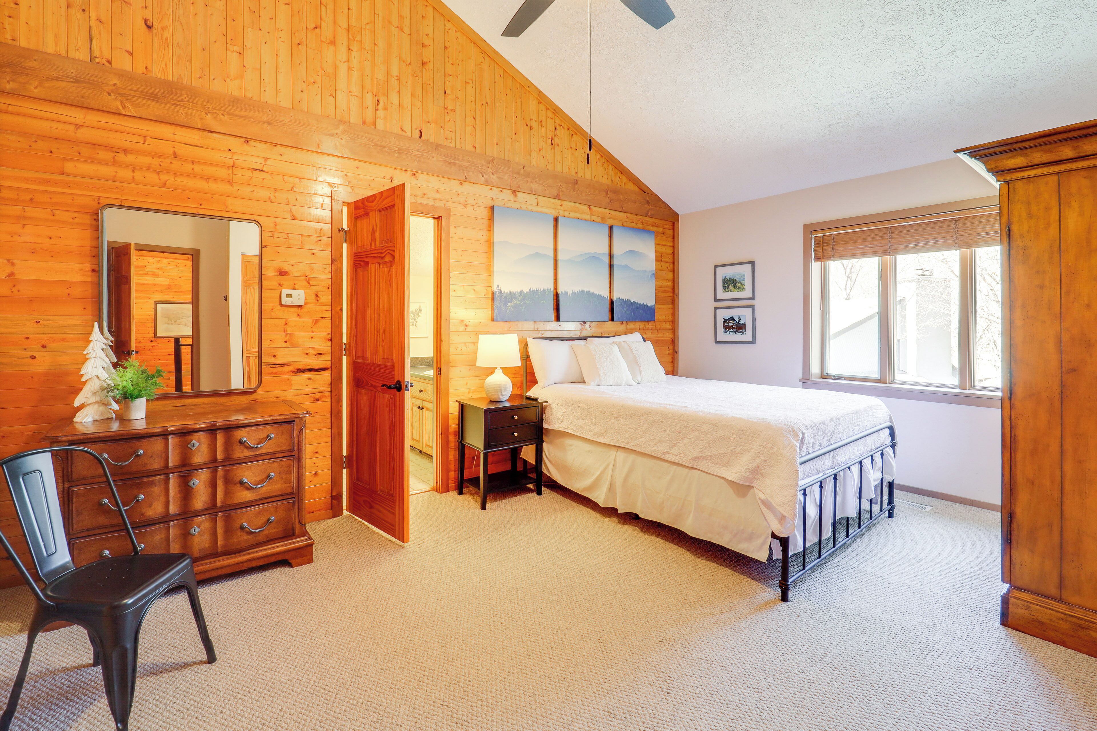 A bedroom with a wooden ceiling, a bed with white bedding, a dresser, a chair, and a nightstand with a lamp.