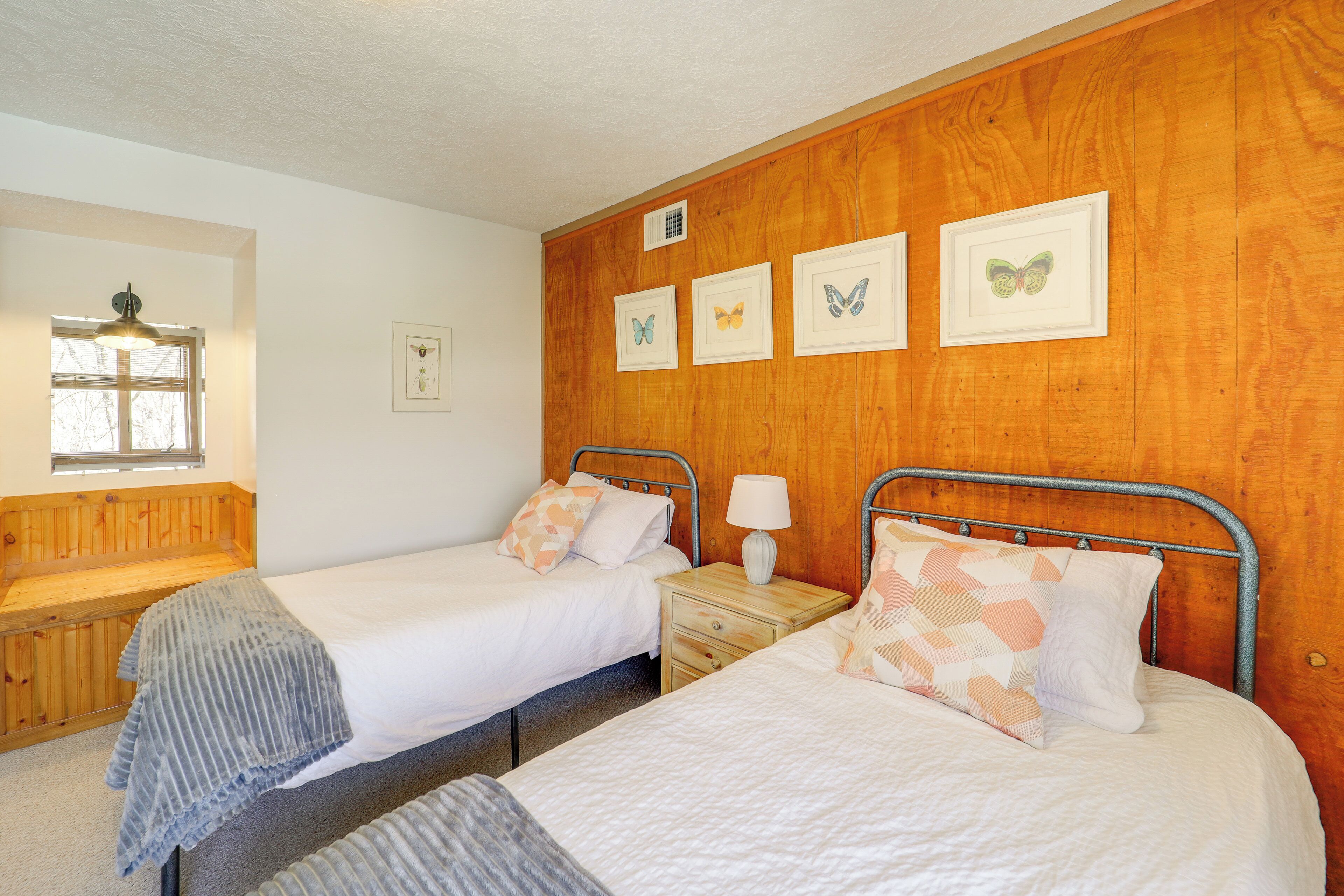Ein Zimmer mit zwei Betten, Holzwandverkleidung, einem Fenster mit Jalousien und gerahmten Schmetterlingsbildern an der Wand.