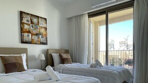 A hotel room with two beds, a large window with curtains, a ceiling light, and a wall-mounted artwork. - Celeste by the Sea: Summer Chill, Pool, Beach 2BR. (Paphos)
