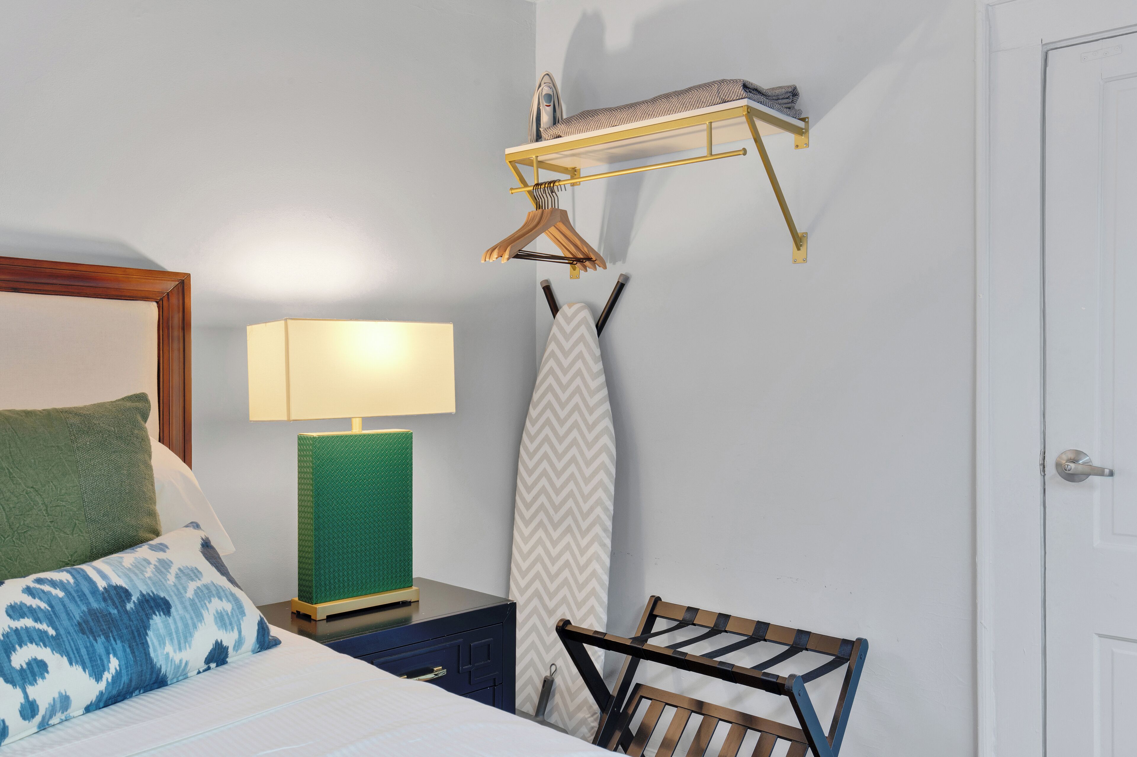 A neatly arranged bedroom with a bed, a bedside lamp, a hanging clothes rack, and a wall-mounted ironing board.