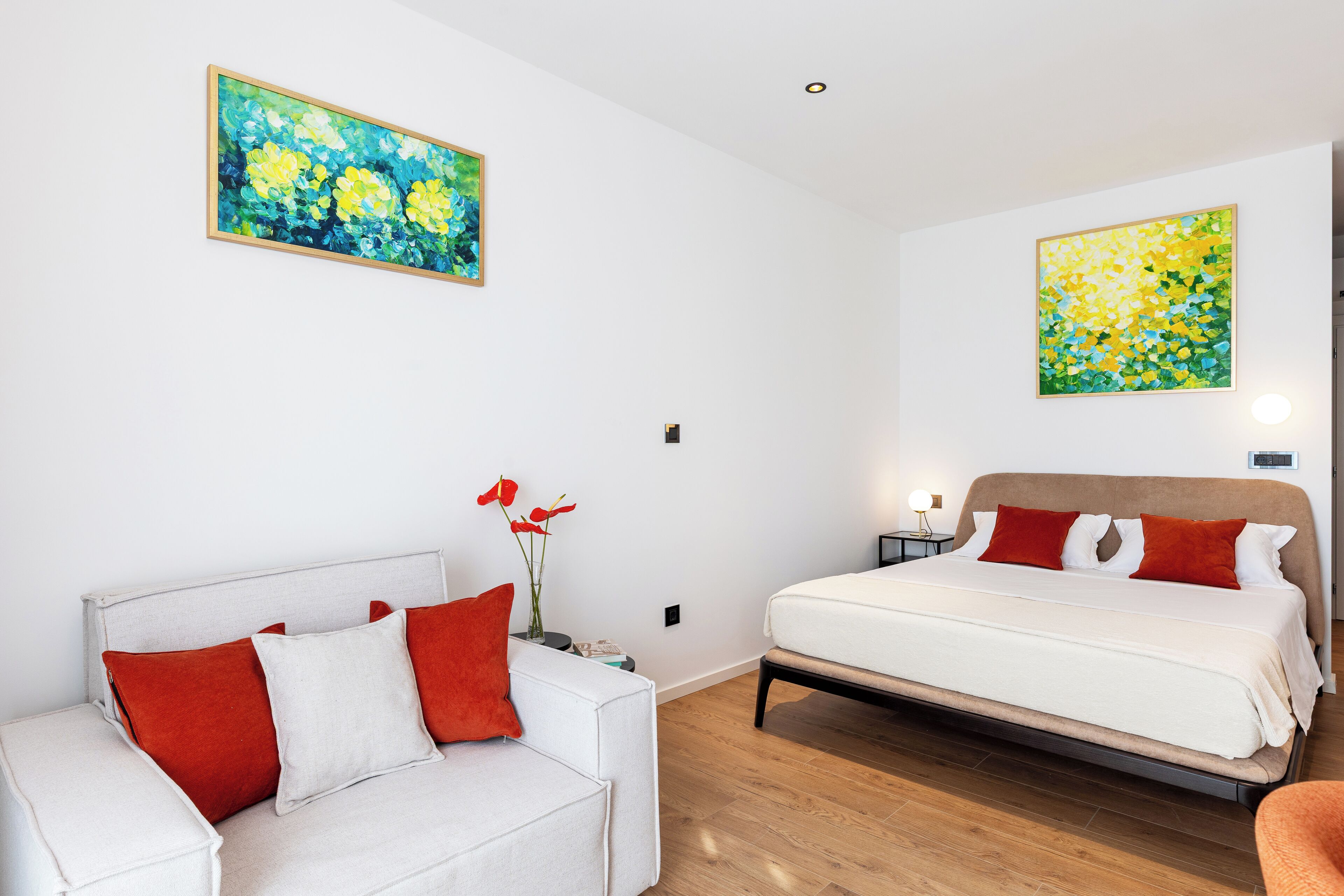 A modern bedroom with a white bed, red and white pillows, a white sofa, and two paintings on the walls.