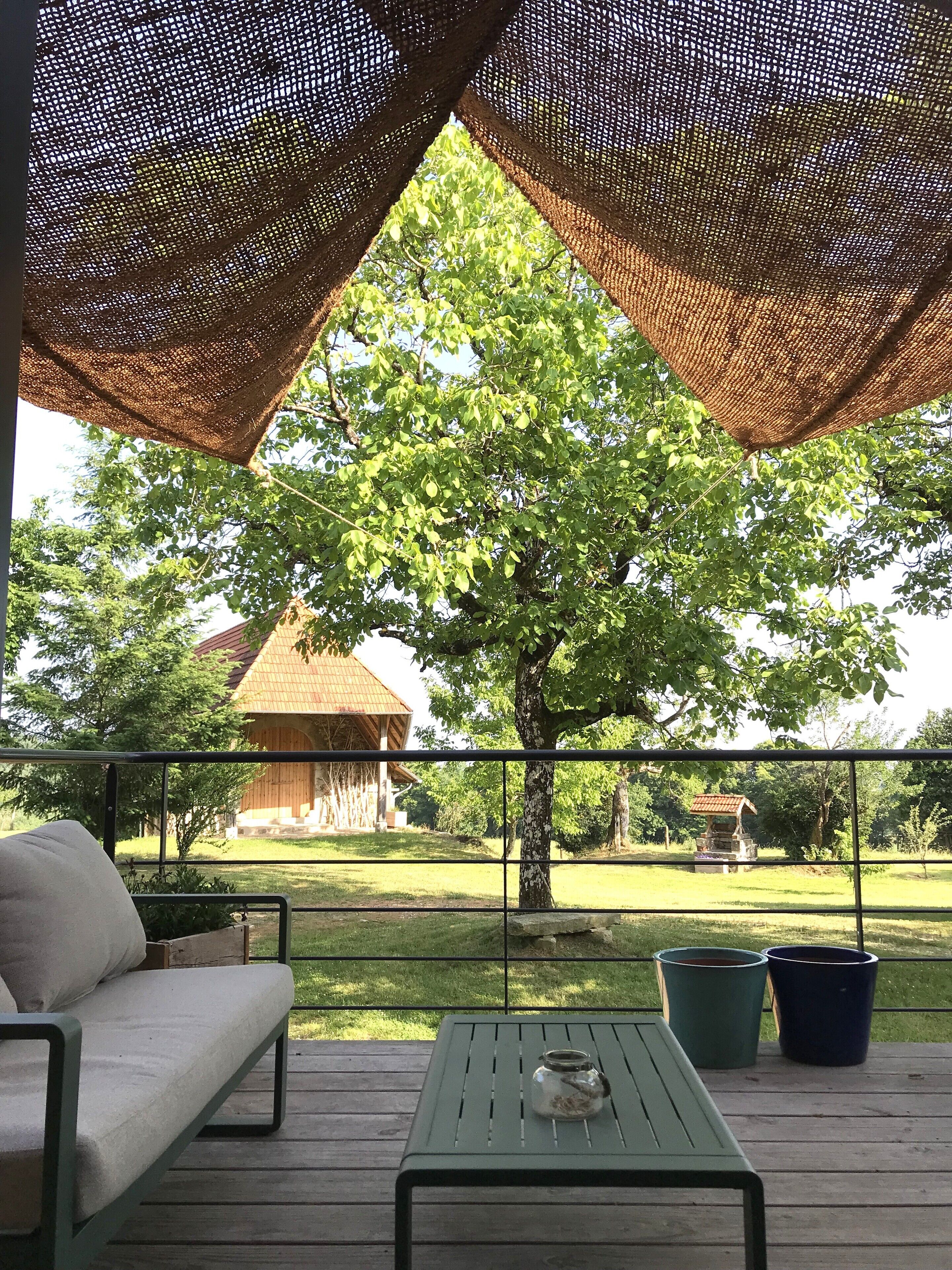 A covered outdoor seating area with a table and chairs, a view of a tree, and a building in the background.