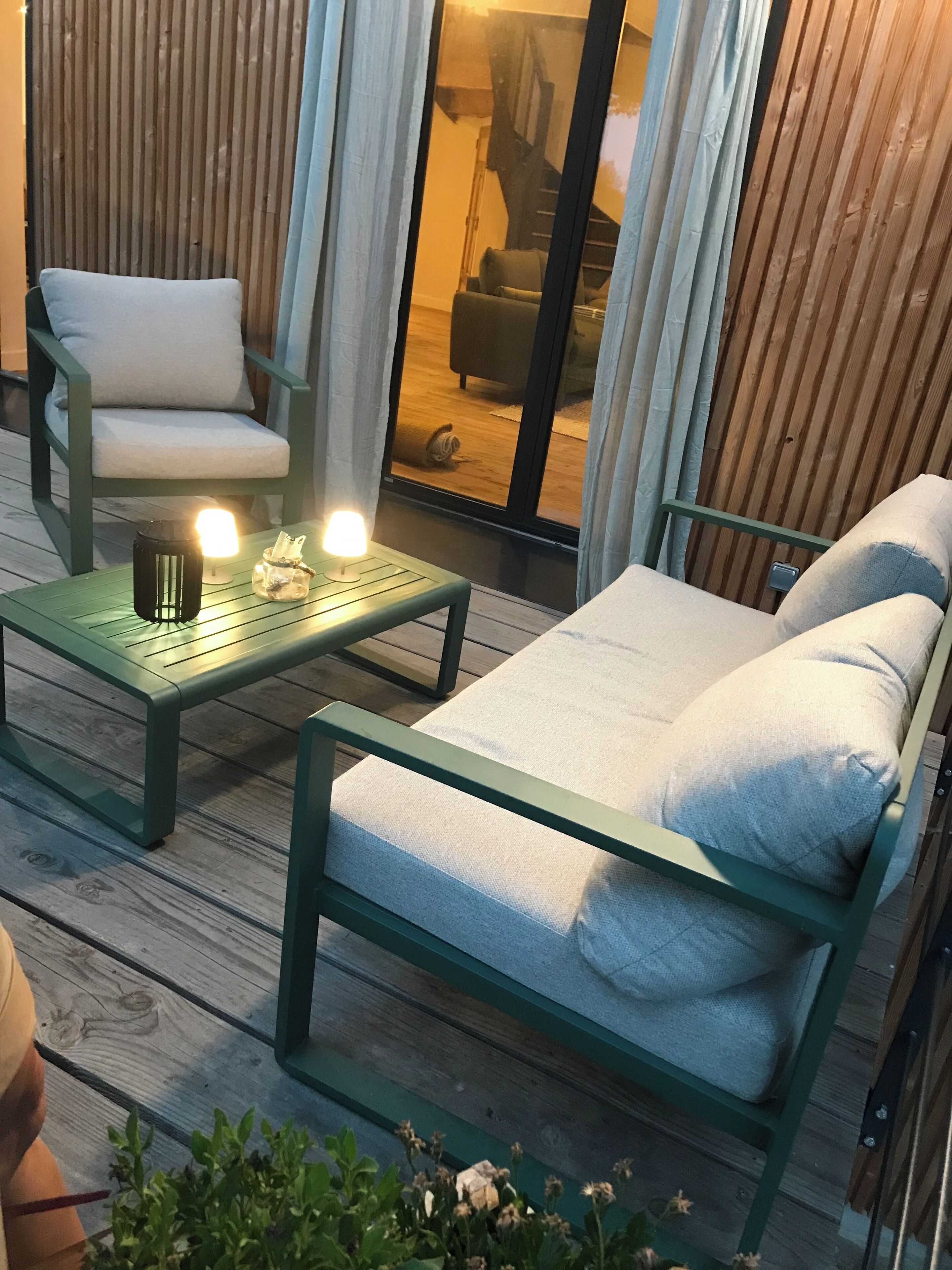 Un espace extérieur accueillant avec un banc en métal vert et deux fauteuils rembourrés. On y trouve une table avec une bougie et un bocal rempli d’en-cas.