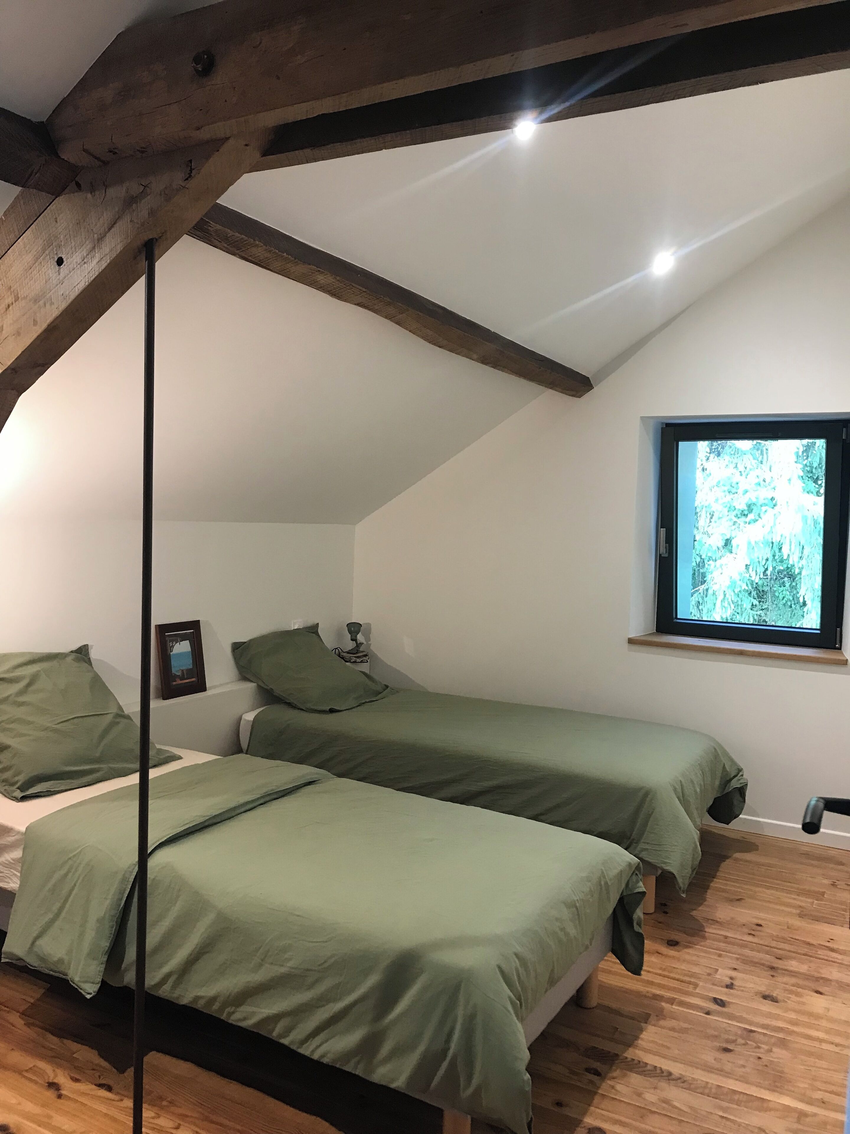 Un dormitori amb dos llits, un sostre de fusta, una finestra amb vista a arbres i una tauleta de nit amb una làmpada.
